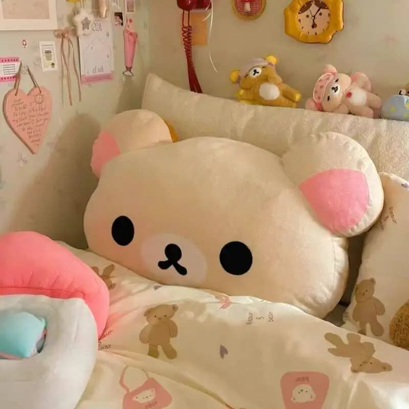 Almofada de pelúcia Rilakkuma, personagem de pelúcia, olhos e nariz pretos, orelhas rosa, corpo bege, fundo rosa e branco.