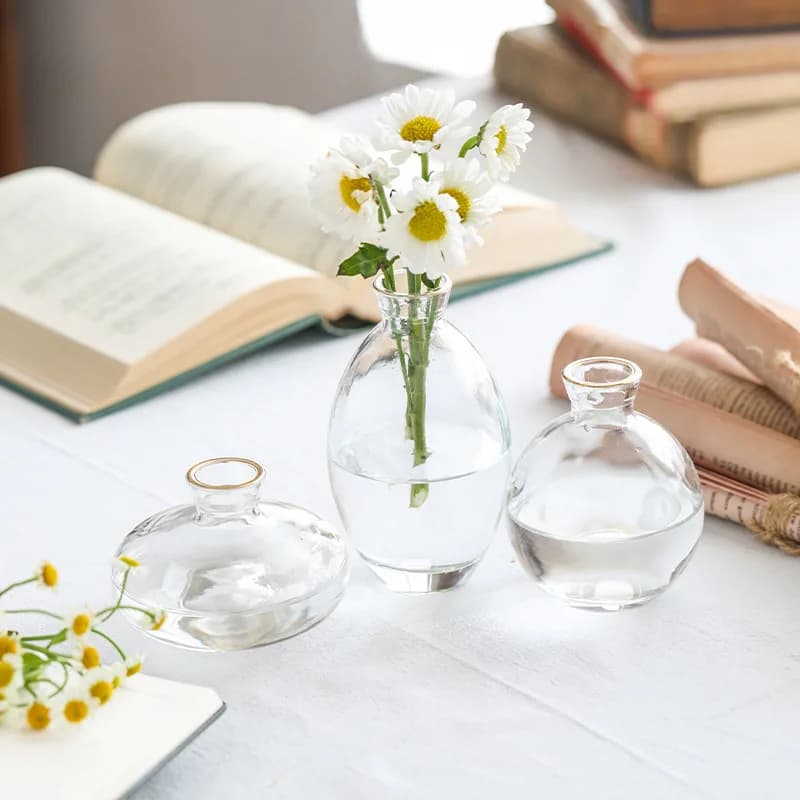 Trê vasos de vidro transparente com flores brancas e amarelas em uma mesa branca