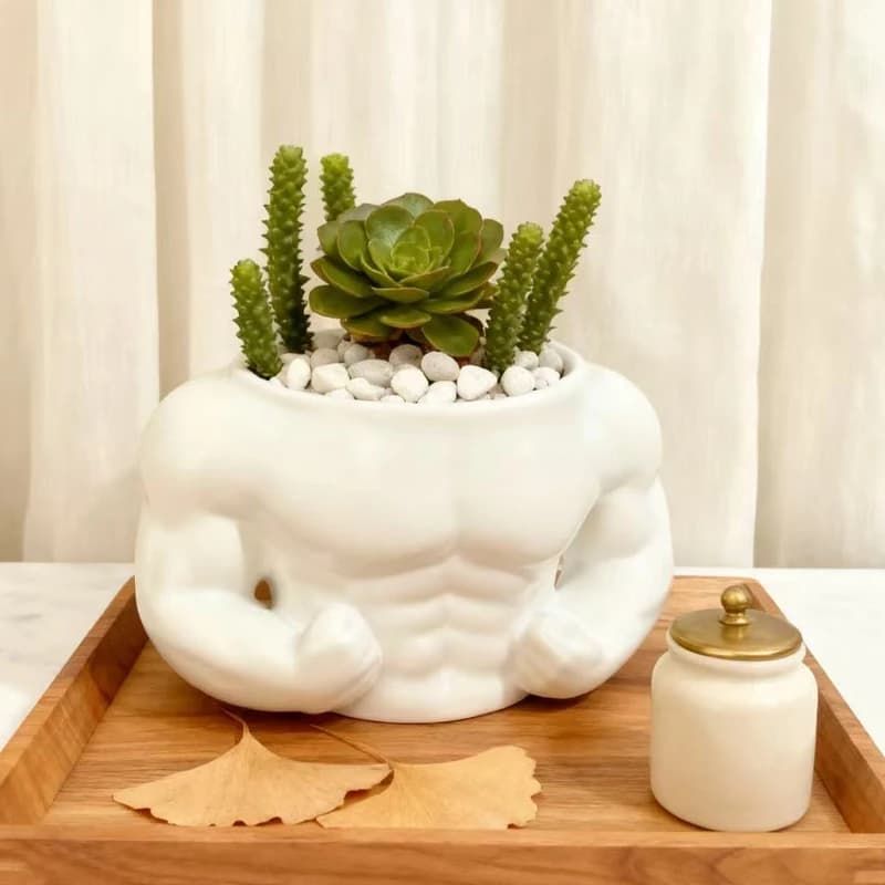 Vaso decorativo em forma de homem musculoso com plantas verdes e cactos, em um suporte de madeira com folhas de ginkgo.