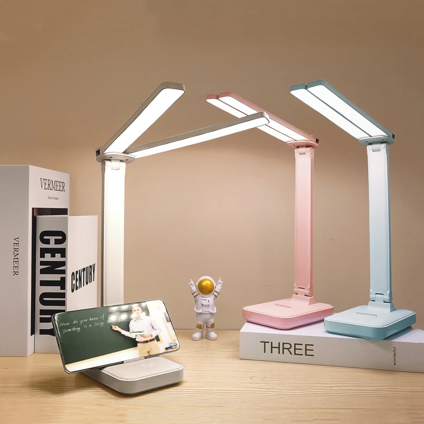 Luminária de mesa USB dobrável em três cores, iluminando um smartphone sobre livros em uma mesa de madeira.