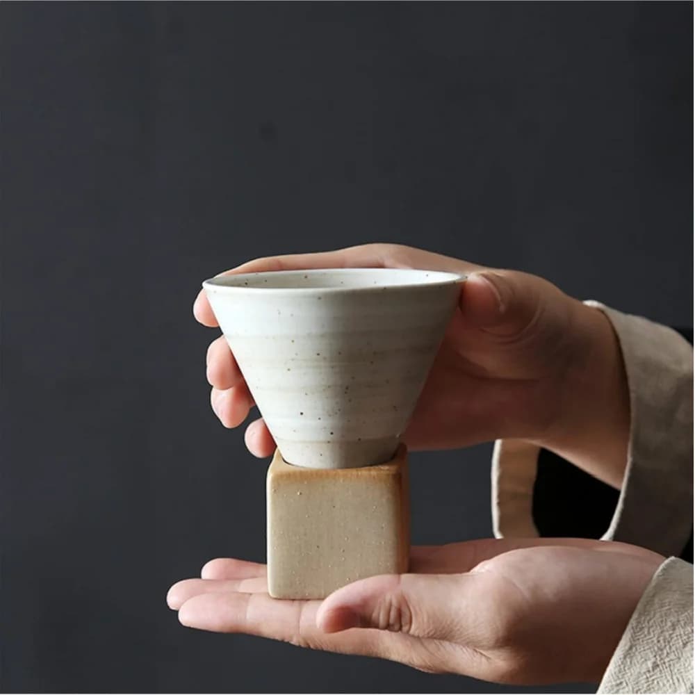 Caneca de café branca com textura e base quadrada bege segurada por duas mãos em fundo escuro