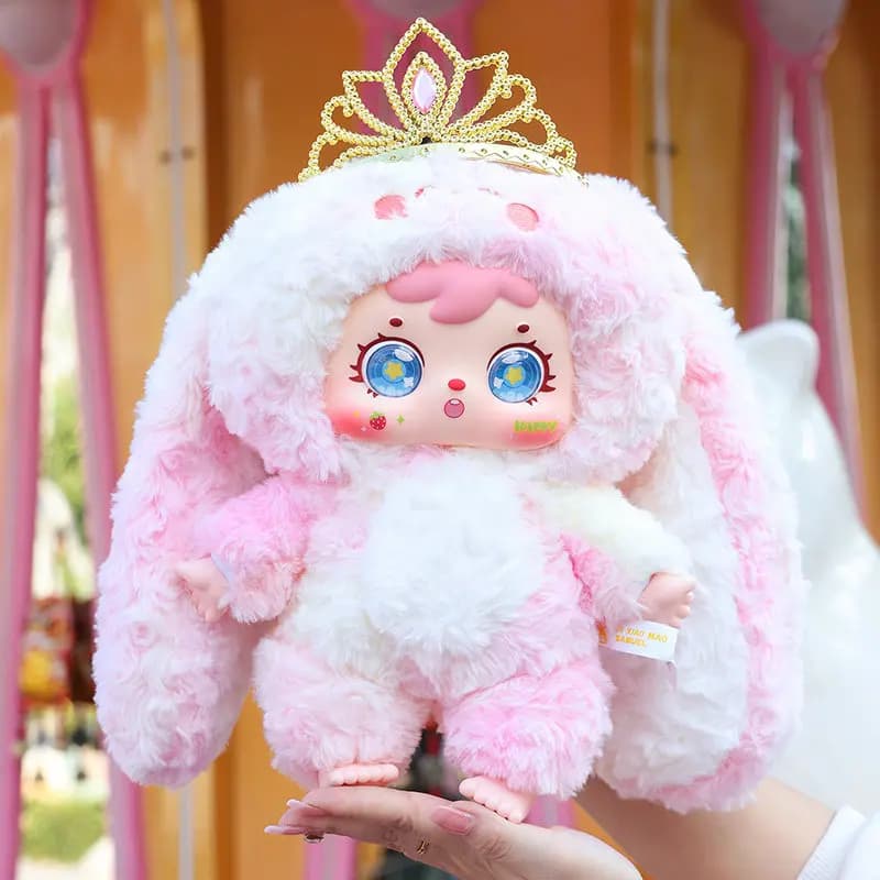 Boneca de pelúcia rosa e branca com coroa dourada, olhos azuis e detalhes coloridos no rosto.