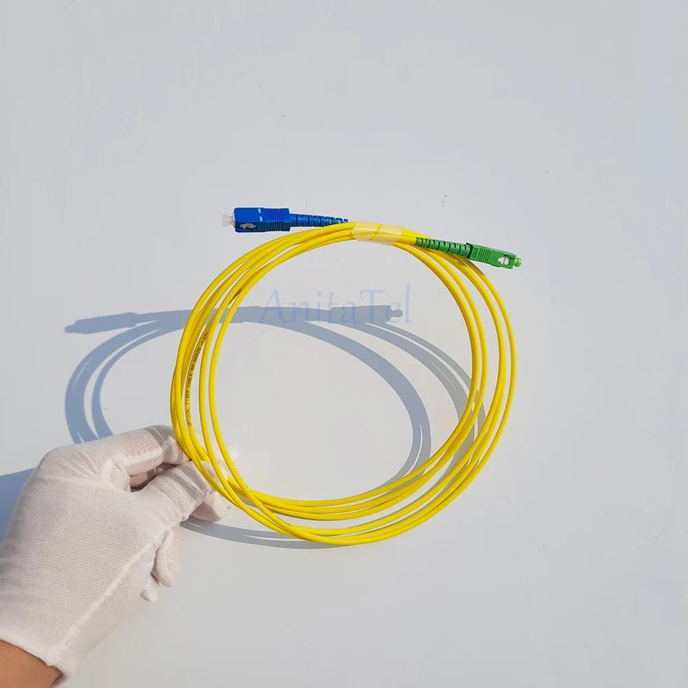 Mão com luva segurando cabo de remendo fibra óptica monomodo amarelo com conectores SC/APC verde e SC/UPC azul em fundo branco