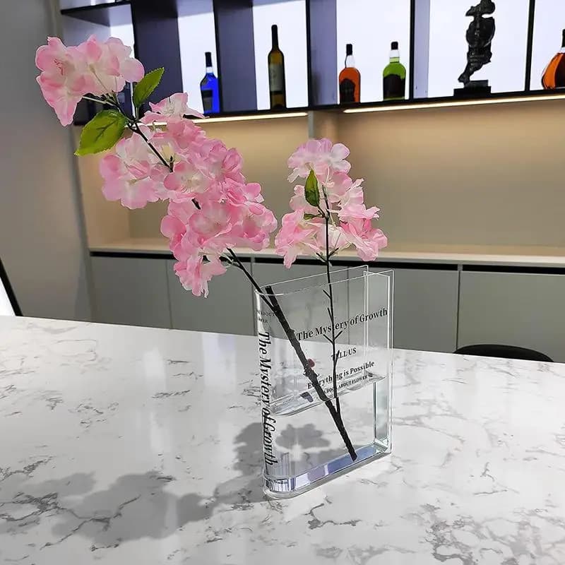 Vaso decorativo acrílico transparente formato livro com flores rosas sobre mesa branca