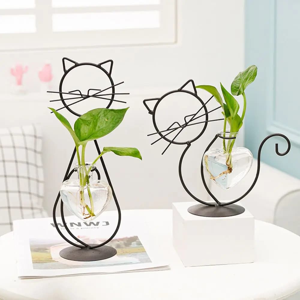 Vaso hidropônico em forma de coração com gato, com plantas verdes, sobre uma mesa branca.