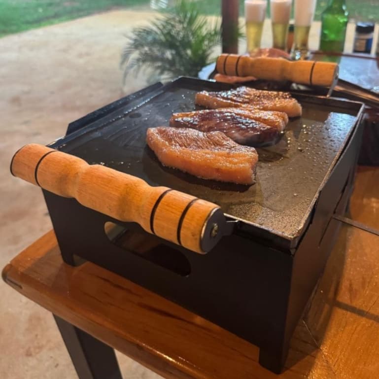 Churrasqueira portátil com chapa de ferro e alça de madeira, grelhando carne em uma mesa externa