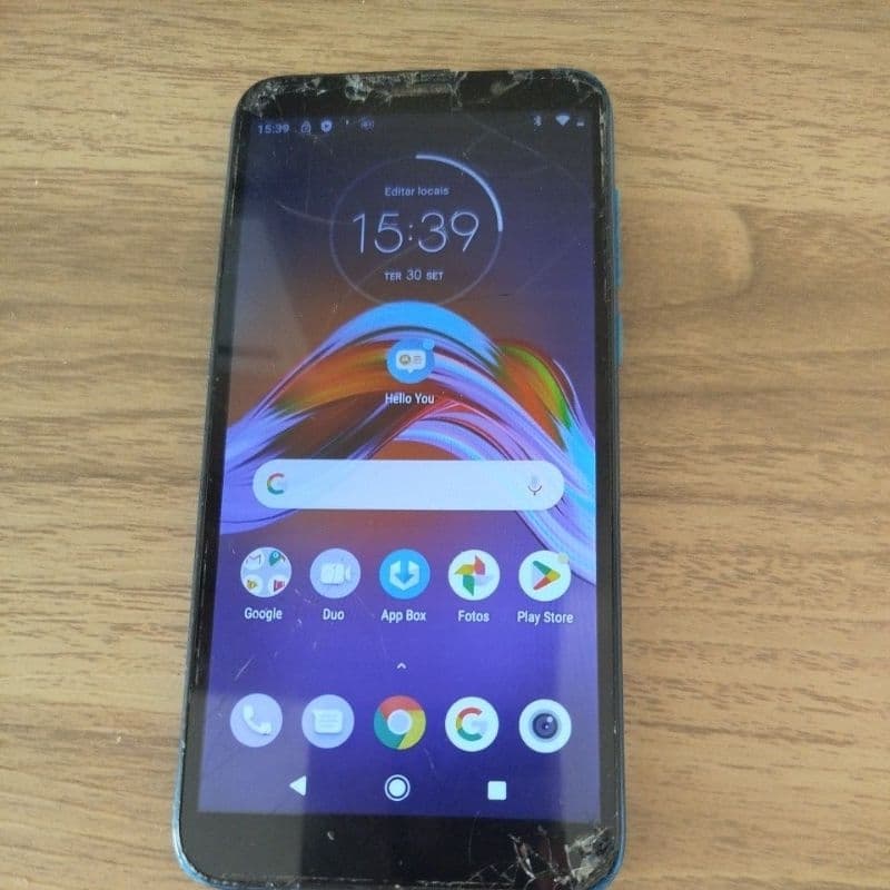 Celular Moto E6 Play quebrado com tela mostrando a hora 15:39 sobre fundo de madeira