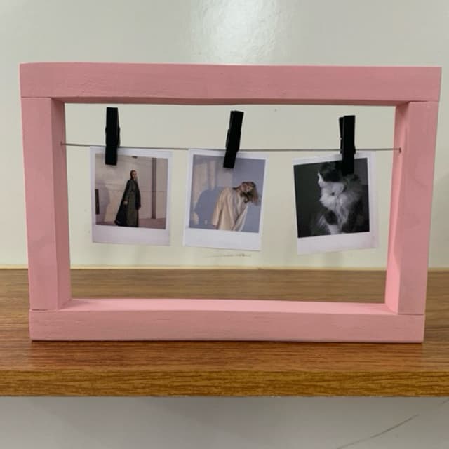 Porta retrato rosa com três fotos polaroid penduradas por clipes, sobre uma superfície de madeira.