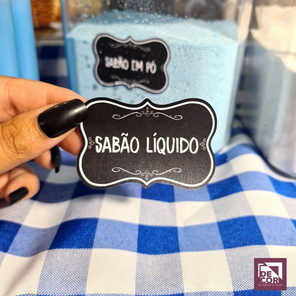 Adesivo preto com bordas douradas e texto "SABÃO LÍQUIDO" sobre toalha xadrez.
