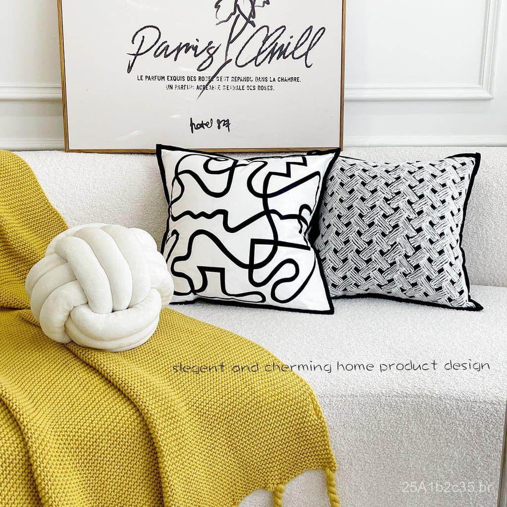 Almofada de sofá em veludo com design abstrato preto e branco, sofá branco, manta amarela, parede branca com quadro dourado.