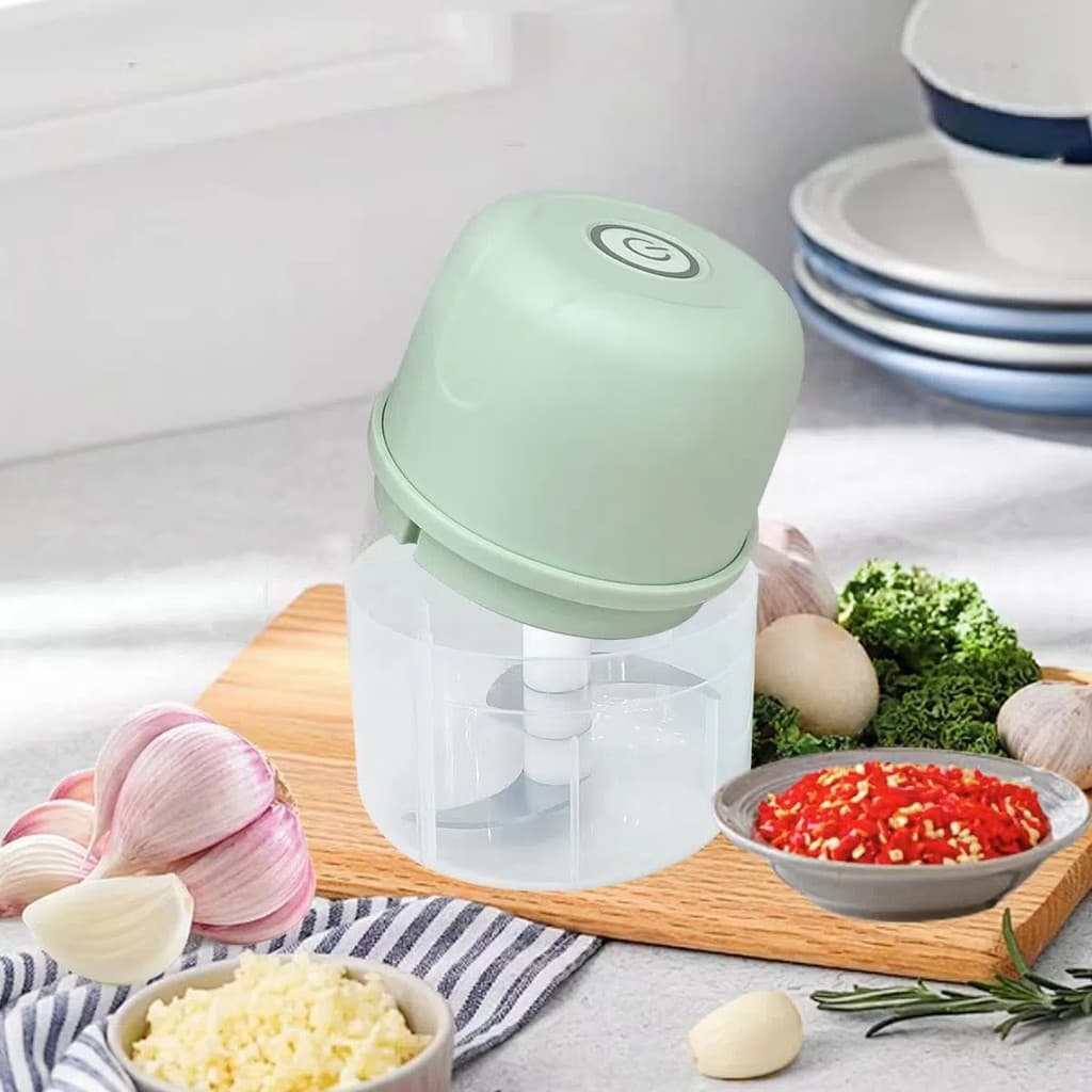 Triturador de alho elétrico pequeno em verde, sobre uma tábua de corte com legumes e temperos.