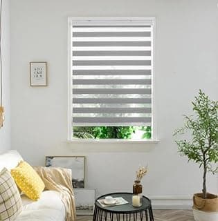 Persiana rolo dupla cinza instalada em uma janela de fundo branco, mostrando uma paisagem verde ao fundo, em uma sala minimalista.