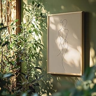 Quadro decorativo minimalista com figura feminina abstrata em preto sobre fundo bege, pendurado em parede verde com plantas.