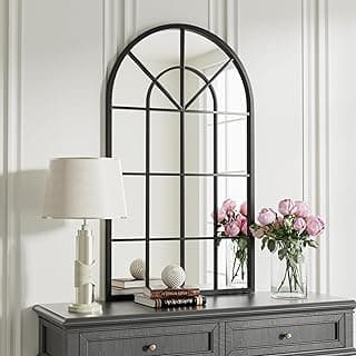 Espelho de parede decorativo com arco preto montado na parede branca, refletindo uma lâmpada, flores e livros