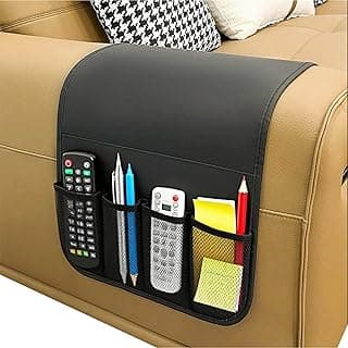 Organizador de apoio de braço de sofá preto com cinco bolsos, contendo controles remotos, canetas e notas adesivas, sobre um sofá bege com almofadas decorativas.