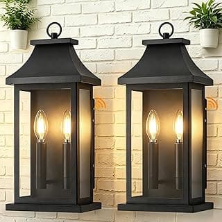 Luminárias de parede externa pretas com cúpulas abauladas e três lâmpadas de filamento, iluminadas em uma parede de tijolos com plantas verdes ao fundo.