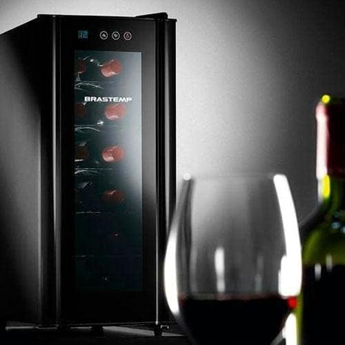 Adega climatizada Brastemp preta com controle digital e porta de vidro transparente, garrafas de vinho dentro, com taça e garrafa de vinho em primeiro plano.