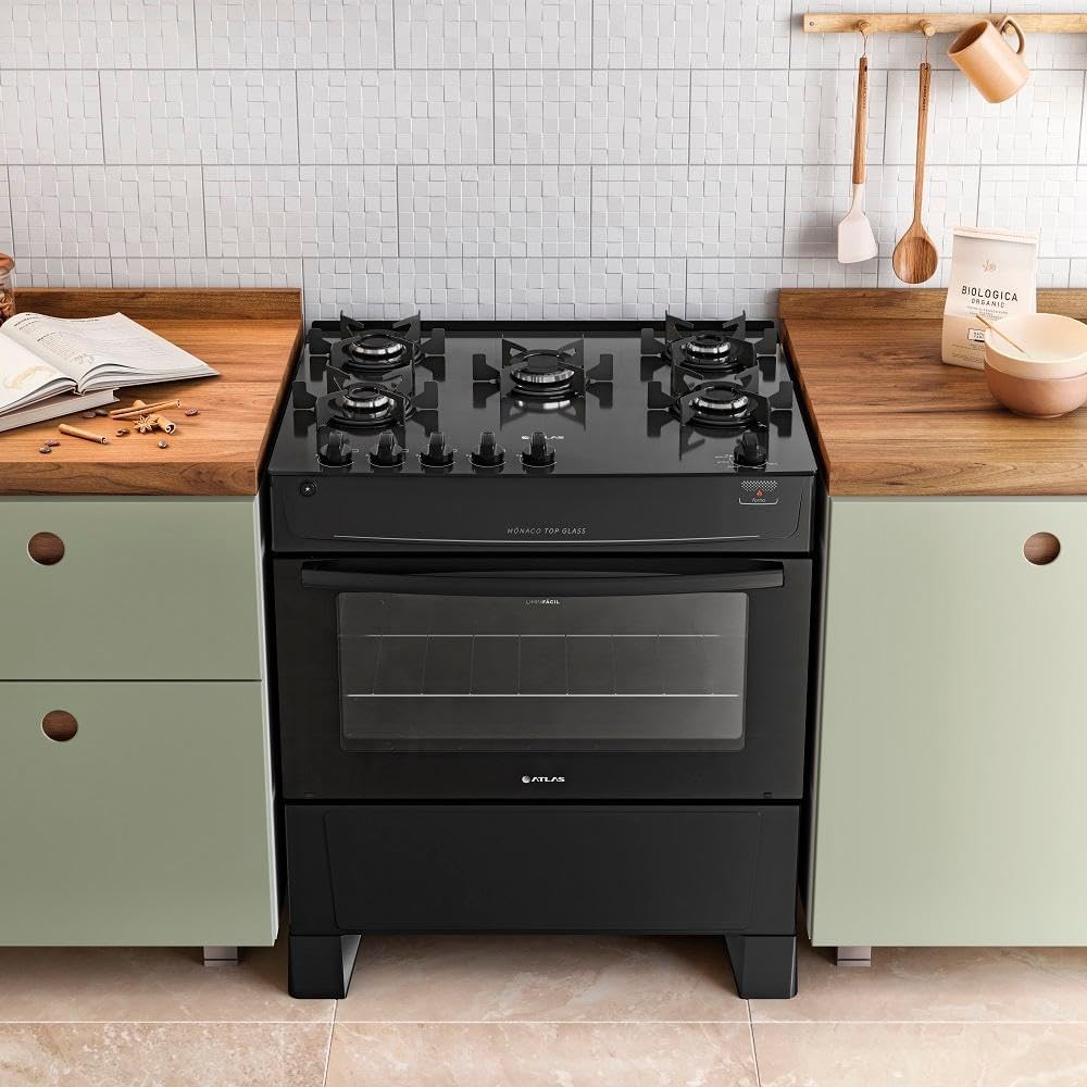 Fogão preto Atlas 5 bocas Mônaco Top Glass instalado em cozinha entre armários verdes e utensílios pendurados